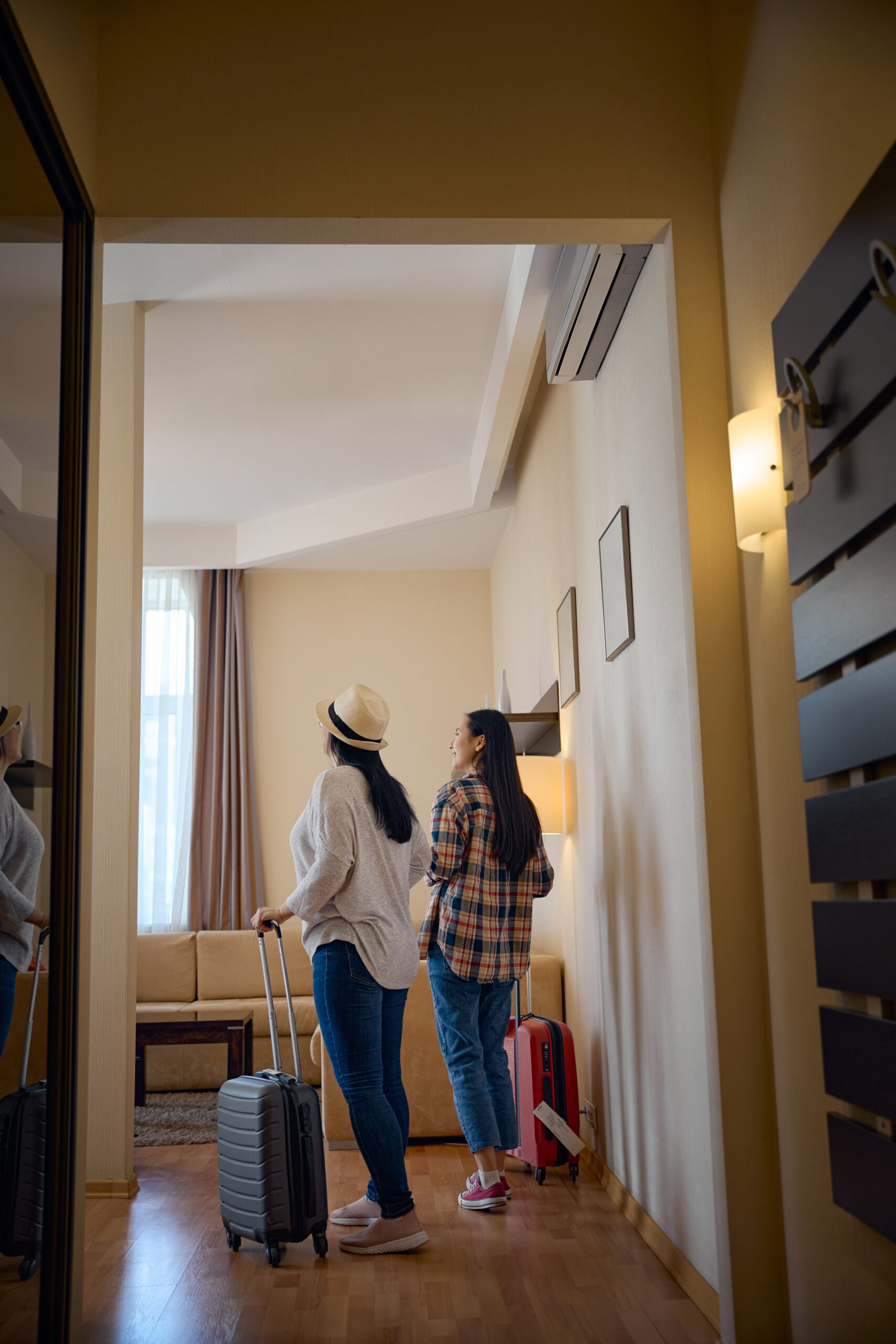 Back view of smiling young Asian woman and her female travel companion standing in middle of hotel room